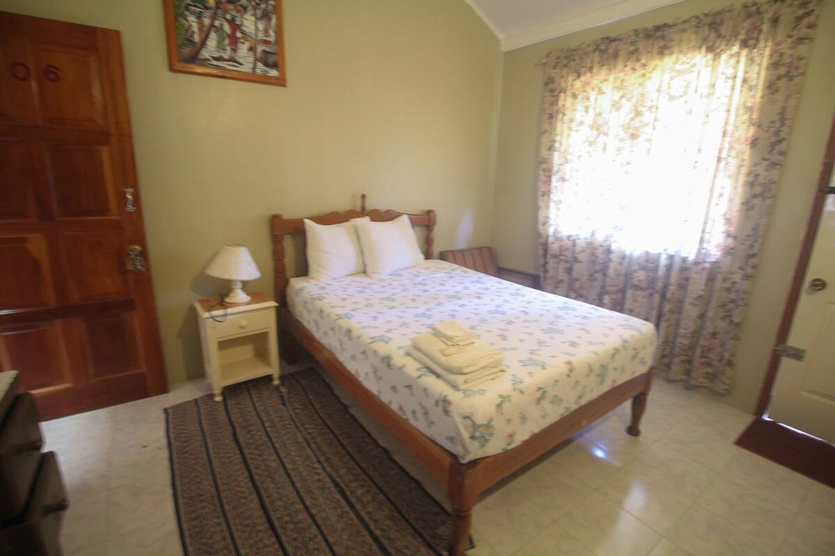 A bedroom with a wooden bed, a bedside table with a lamp, a window with floral curtains, and a door.