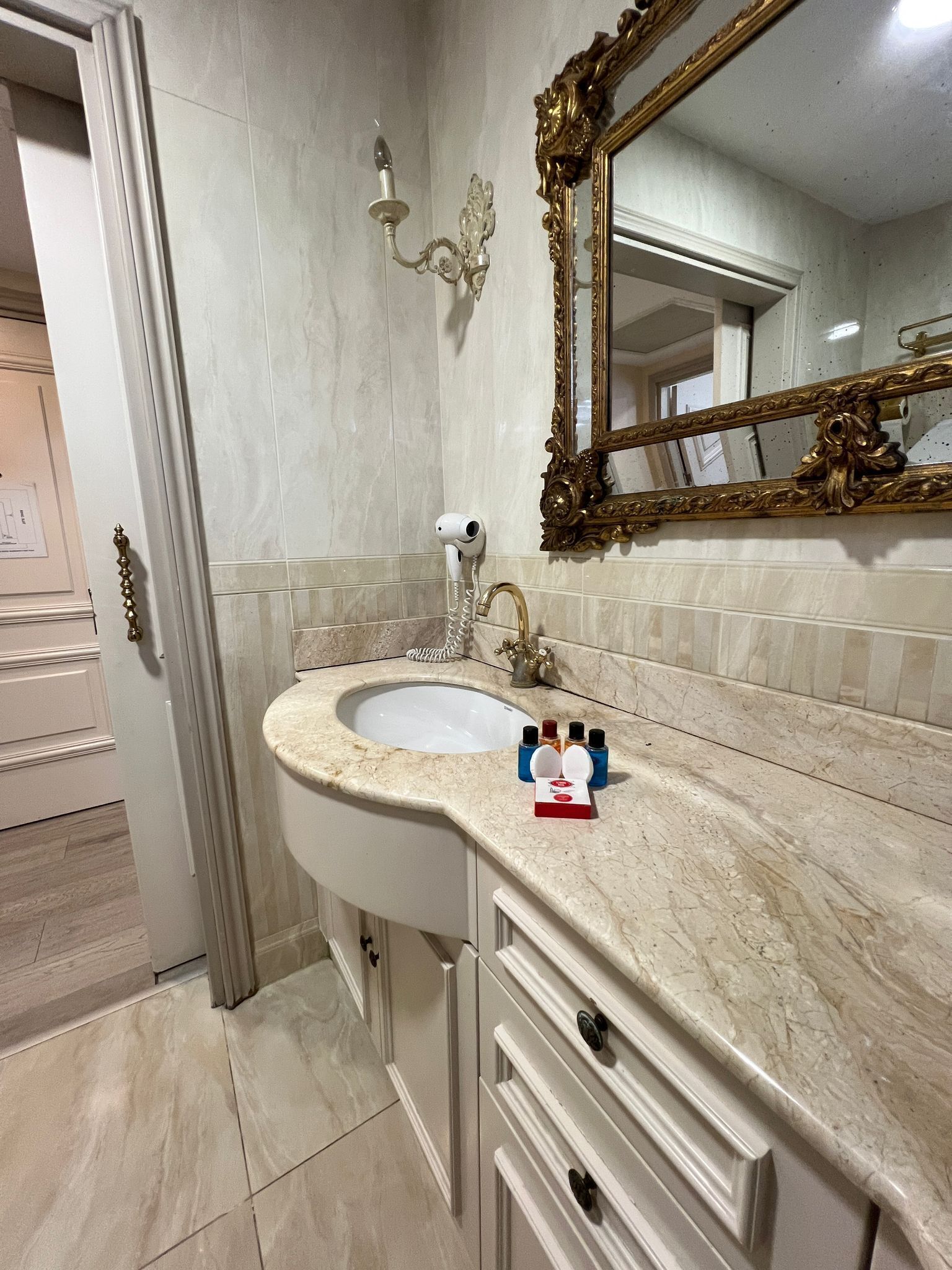 A bathroom with a marble sink, a gold-framed mirror, and a wall-mounted light fixture.