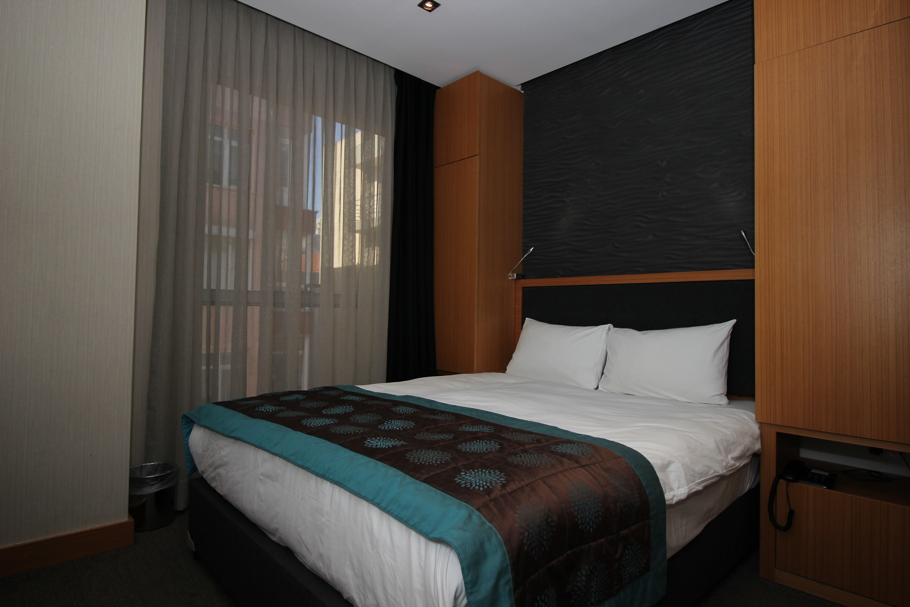 A hotel room with a bed, a wooden headboard, a window with sheer curtains, and a small trash bin.