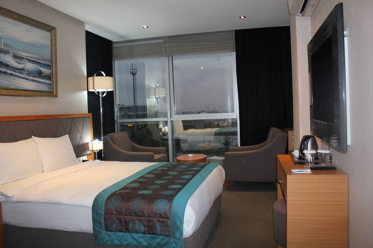 A hotel room with a large bed, a desk, a chair, and a view of the city through a large window.