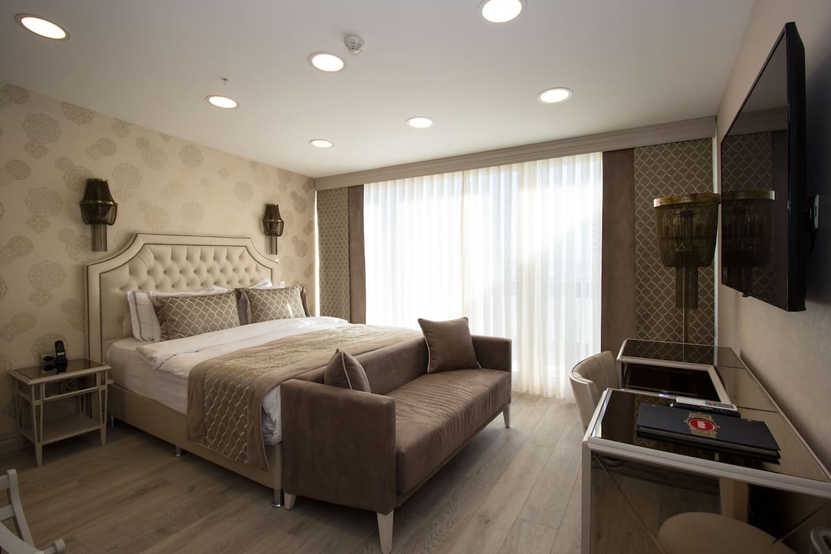 A modern hotel room with a large bed, a sofa, a desk with a laptop, and a TV on the wall.