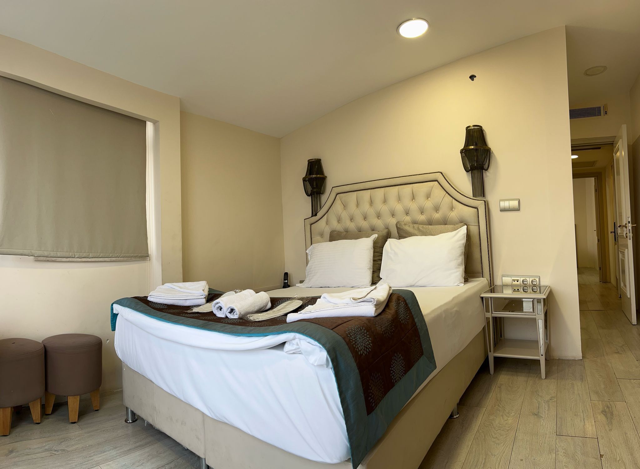 A neatly made bed with a tufted headboard, a bedside table with a lamp, and two ottomans.