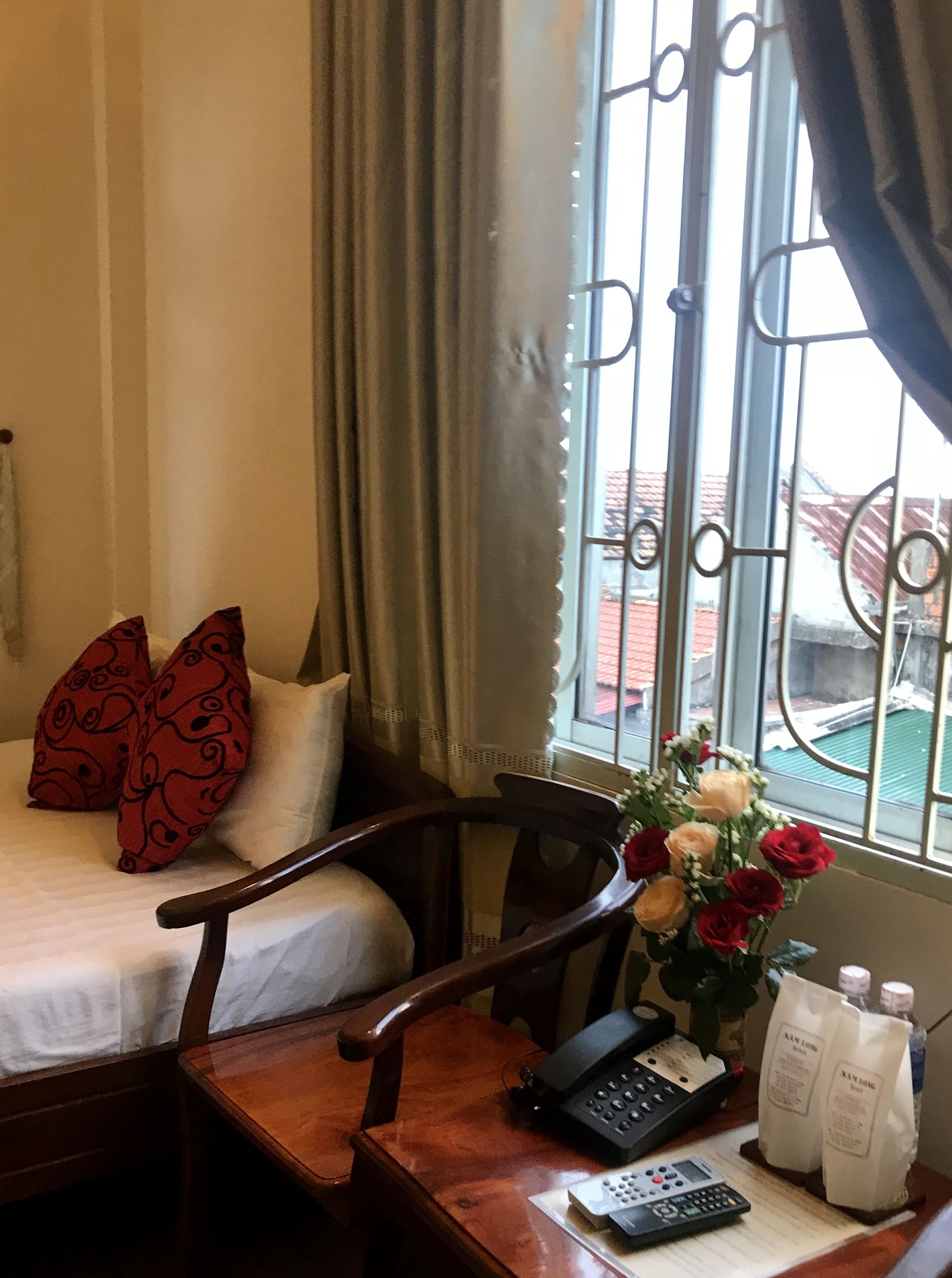 Habitación de hotel con cama, una silla de madera, un escritorio con teléfono y control remoto, un jarrón con flores y una ventana con barrotes.