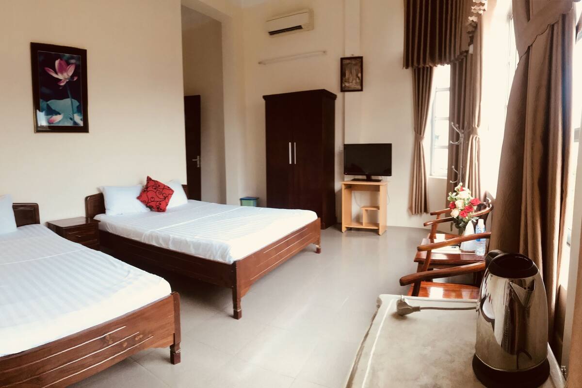 A hotel room with two wooden beds, a TV, a small table with a kettle, and a window with curtains.