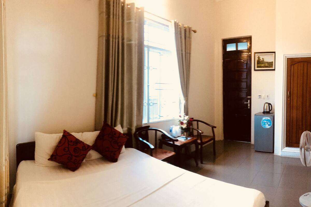 A hotel room with a bed, a desk, a chair, a fan, and a door.