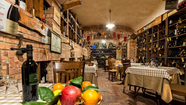 A wine cellar with wooden shelves and tables, featuring a variety of wine bottles and a basket of fruits. - Hotel Ristorante Donato (Calvizzano)
