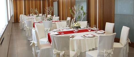 Un salón de banquetes con mesas redondas, sillas blancas y manteles rojos.