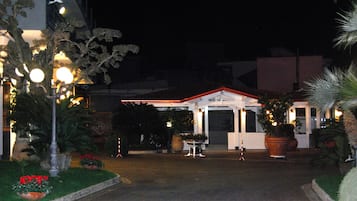 L’entrée d’un hôtel la nuit, avec une façade bien éclairée, un espace d’attente abrité et des plantes en pot.