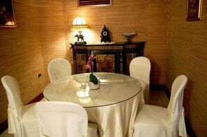A round table with a glass top, surrounded by white chairs, in a room with wooden paneling and a decorative cabinet with a clock and an elephant figurine.