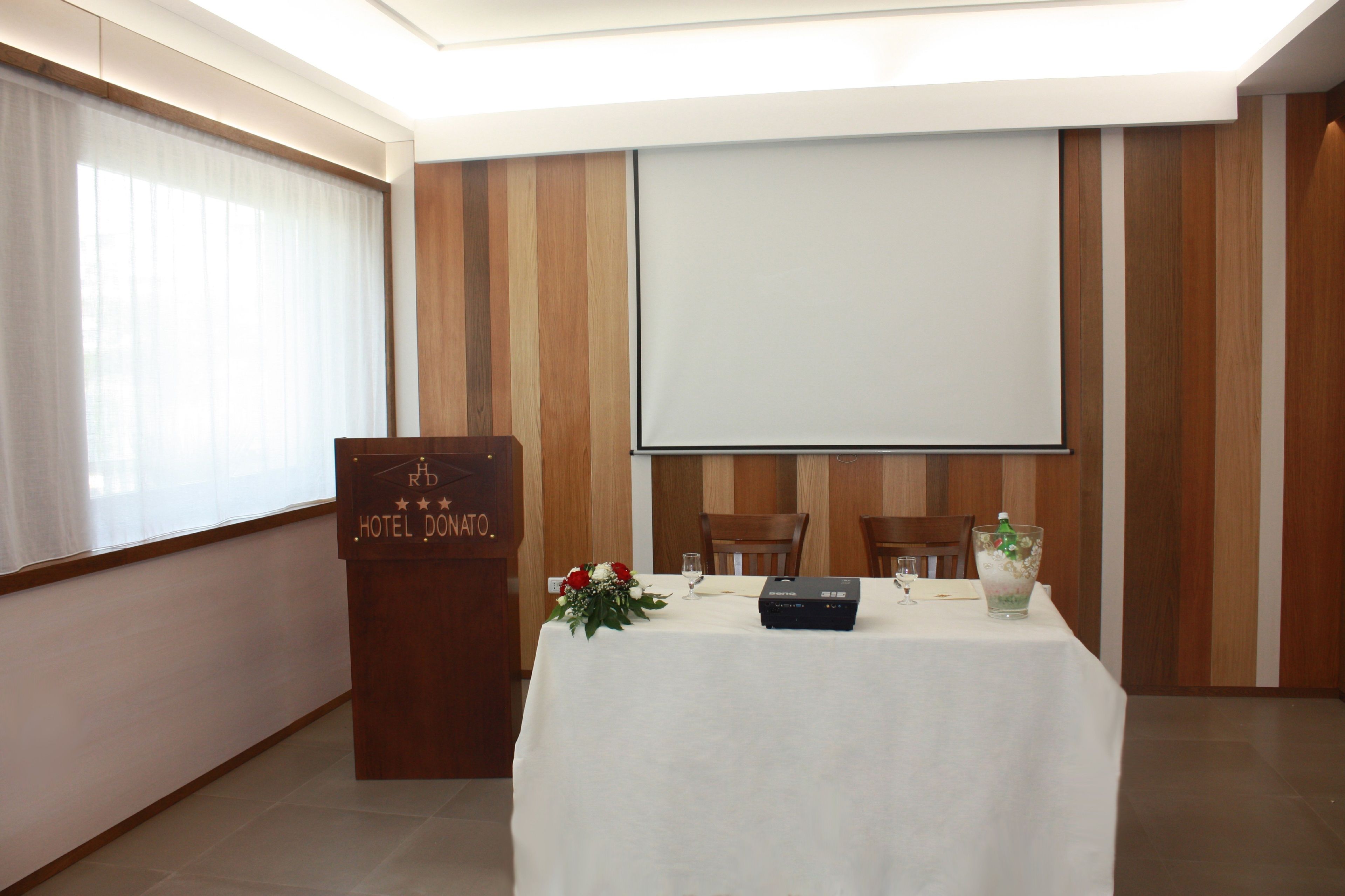 Un salón de conferencias con un atril, una pantalla de proyector, una mesa cubierta con mantel blanco y dos sillas.