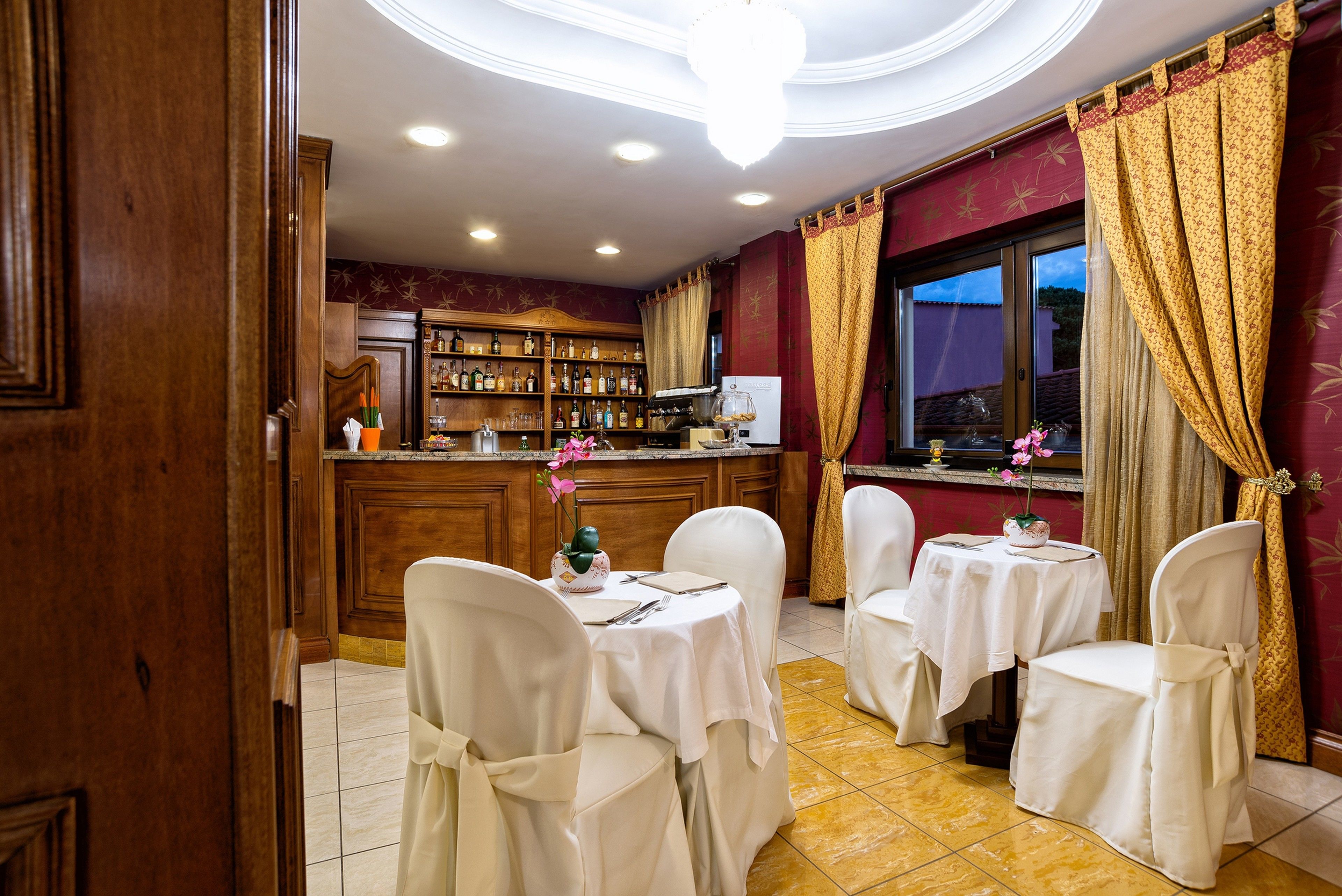 A bar area with wooden cabinets, a counter, and a large window with curtains. There are two round tables with chairs, each covered with a white tablecloth.