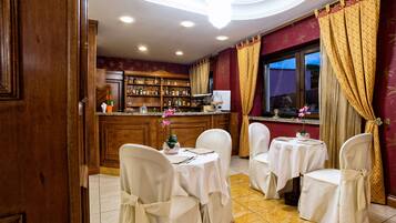 A bar area with wooden cabinets, a counter, and a large window with curtains. There are two round tables with chairs, each covered with a white tablecloth.