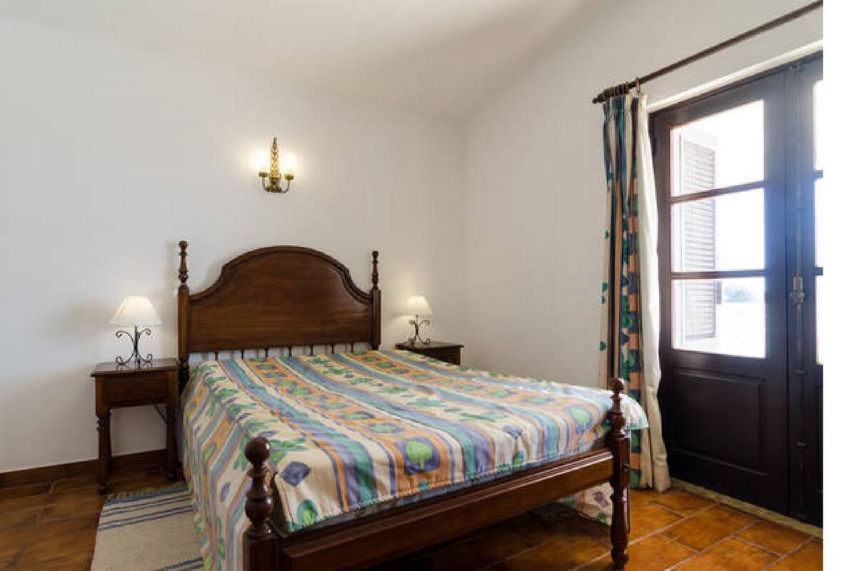 Une chambre à coucher avec un lit en bois, deux tables de chevet avec des lampes, une fenêtre avec des rideaux et une porte.