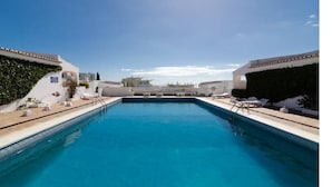 Ein rechteckiger Swimmingpool mit glasklar blauem Wasser, umgeben von einer weißen Poolterrasse. Auf einer Seite stehen Liegestühle und ein kleiner Tisch, auf der anderen eine Hecke.
