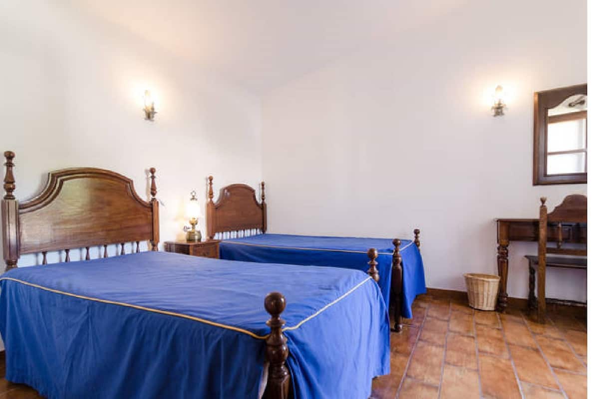 Une chambre avec deux lits en bois, chacun recouvert d’une courtepointe bleue, une table de chevet en bois avec une lampe, une chaise en bois et une petite table en bois.