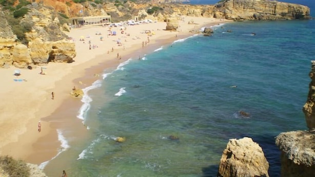 Ein Küstenstrand mit klarem Wasser, felsigen Klippen und einem Gebäude mit Strandliegen.
