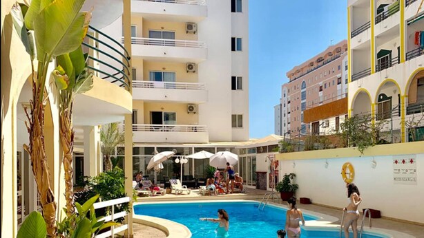 A swimming pool area with people swimming and lounging, surrounded by multi-story buildings with balconies and outdoor seating. - Aparthotel Calema Avenida Jardim (Vila Real Santo Antonio)