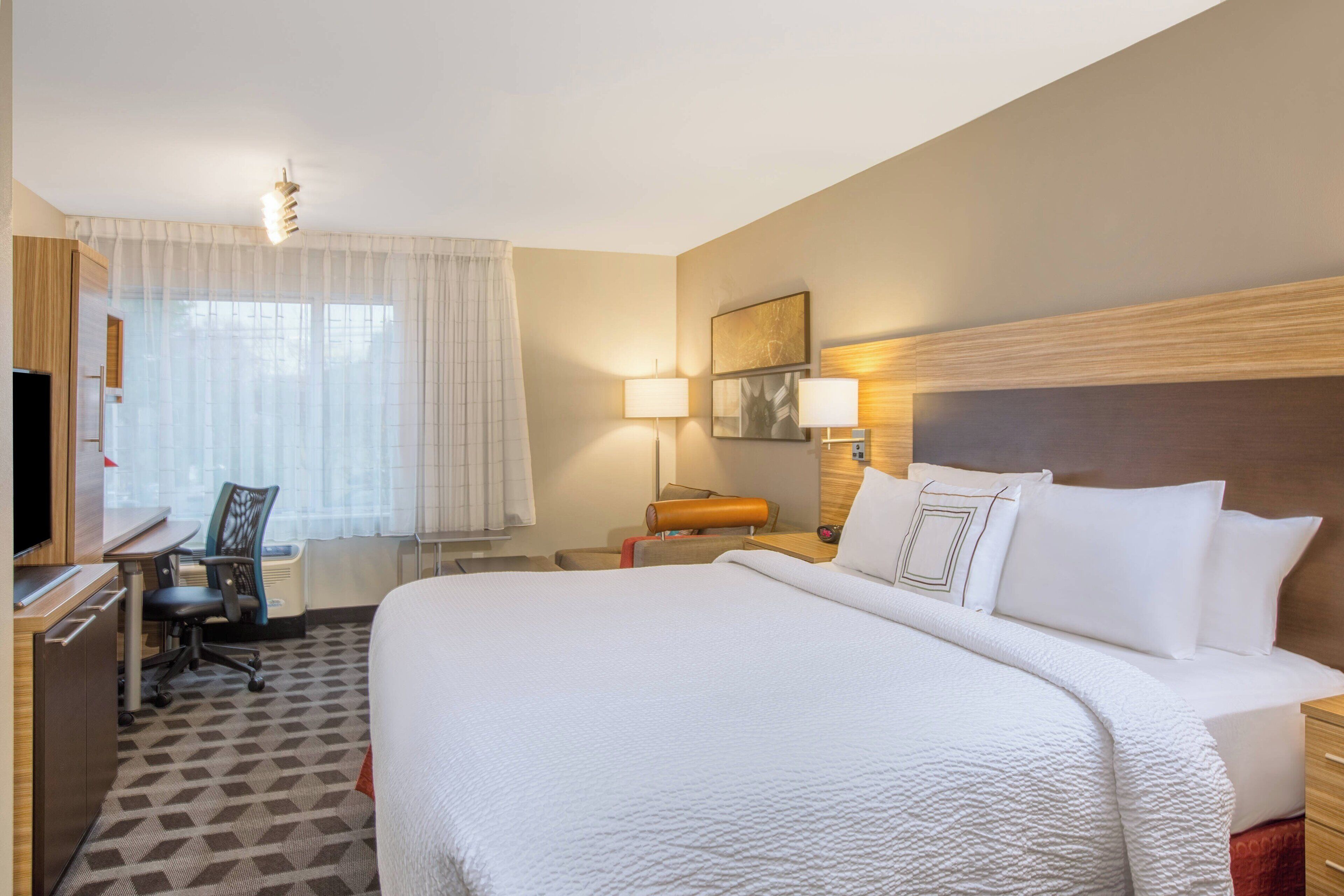 A hotel room with a large bed, a desk with a chair, a television, and a window with sheer curtains.
