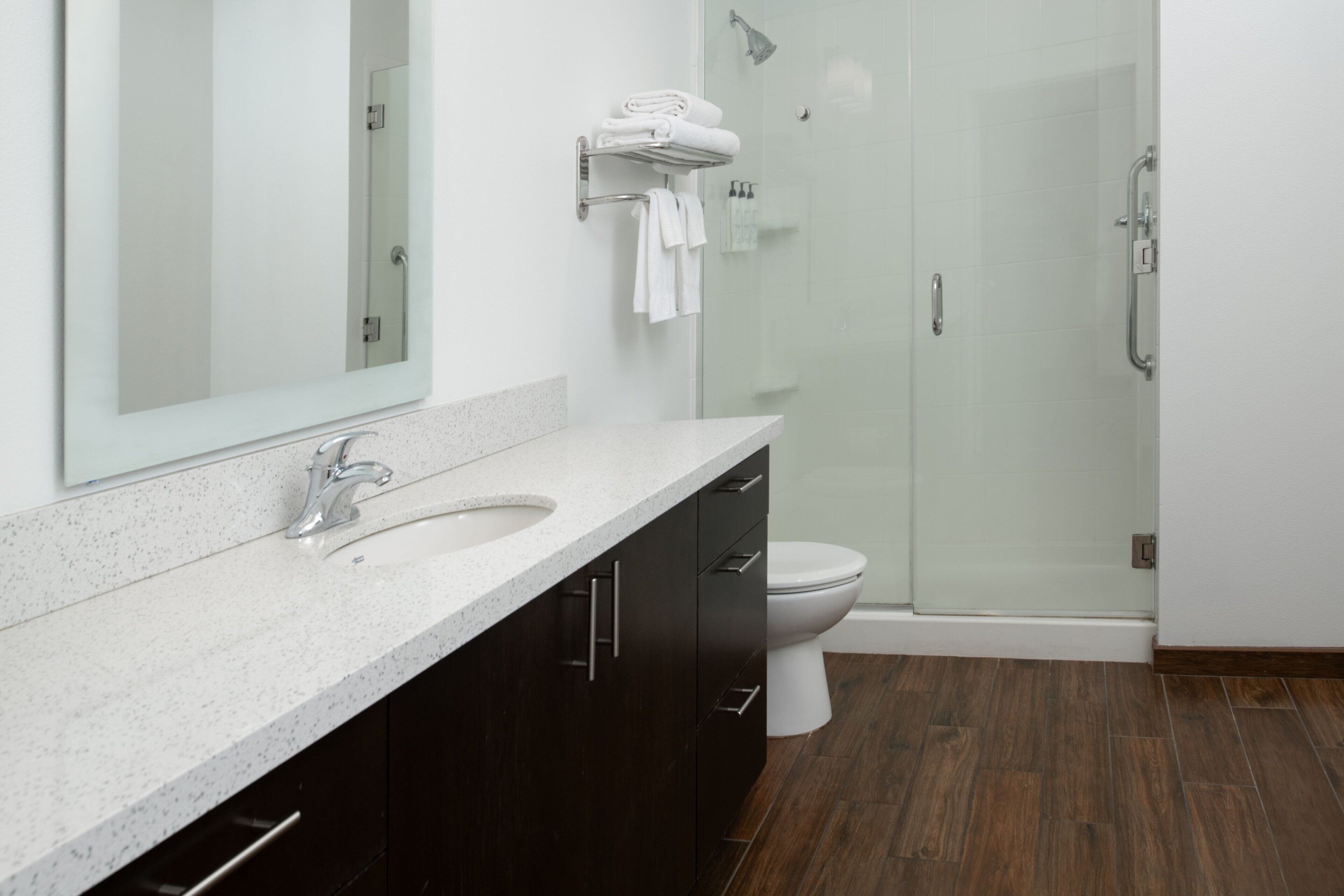 Une salle de bain moderne dotée d’un grand miroir, de deux lavabos, de meubles sombres, de toilettes et d’une douche séparée par des parois en verre.