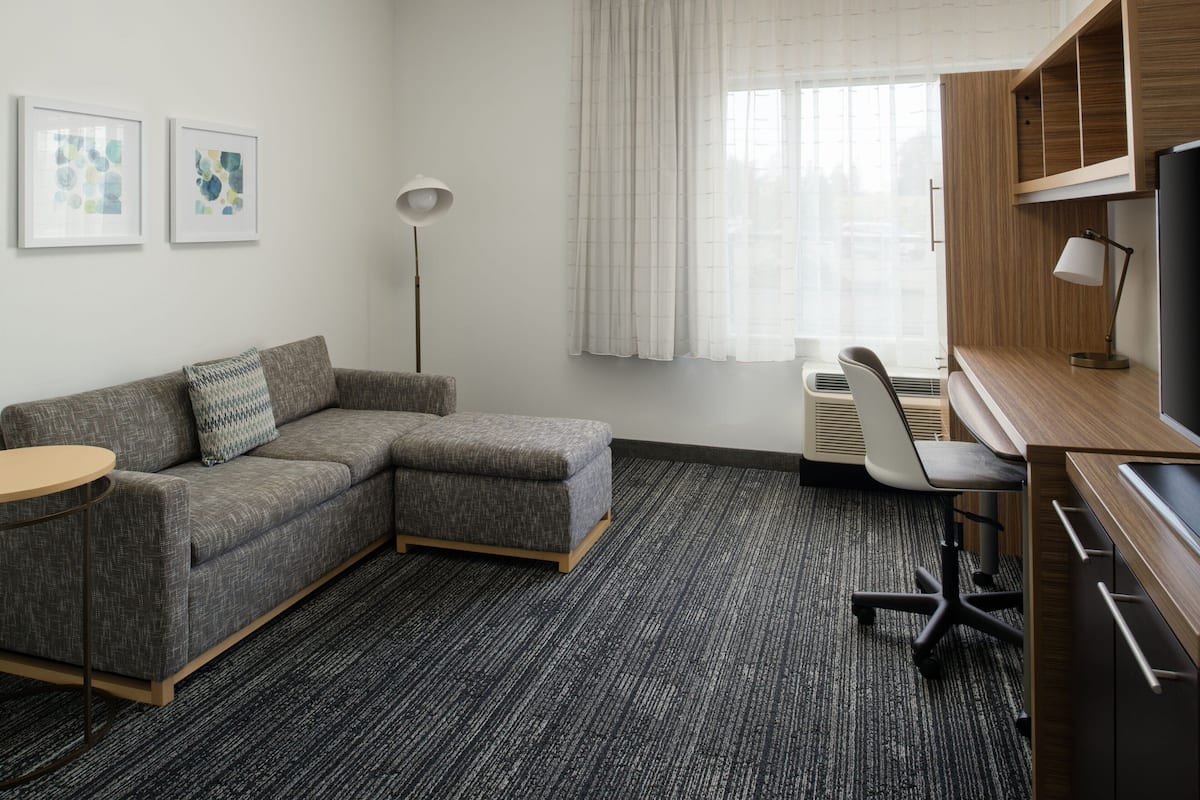 Moderni hotellihuone, jossa on harmaa kulmasohva, pyöreä sivupöytä, työpöytä tuolilla, lattiavalaisin ja suuri ikkuna, jossa on verhot.