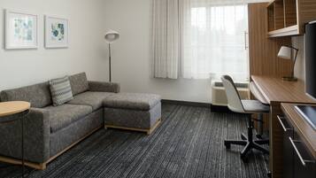 A modern hotel room with a grey sectional sofa, a round side table, a desk with a chair, a standing lamp, and a large window with curtains.