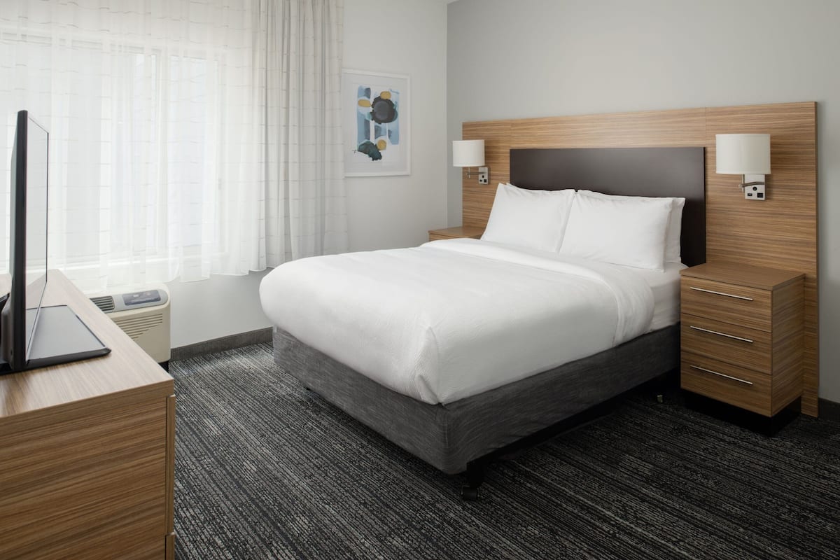 Hotellihuone, jossa on suuri sänky, yöpöydät, työpöytä kannettavalla tietokoneella ja ikkuna, jossa on läpikuultavat verhot.