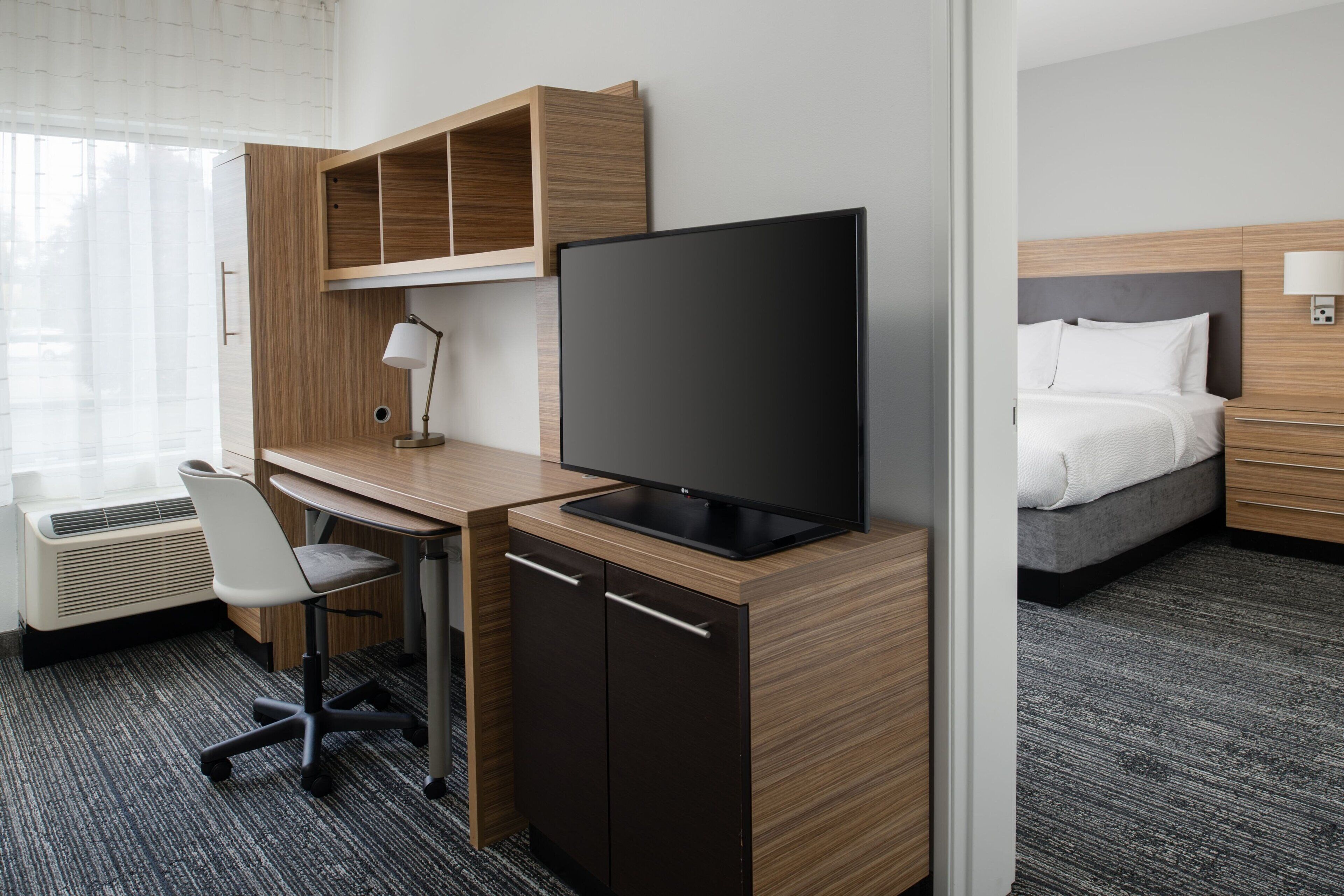 Una habitación de hotel con un escritorio de madera, televisor, cama y ventana con cortinas.
