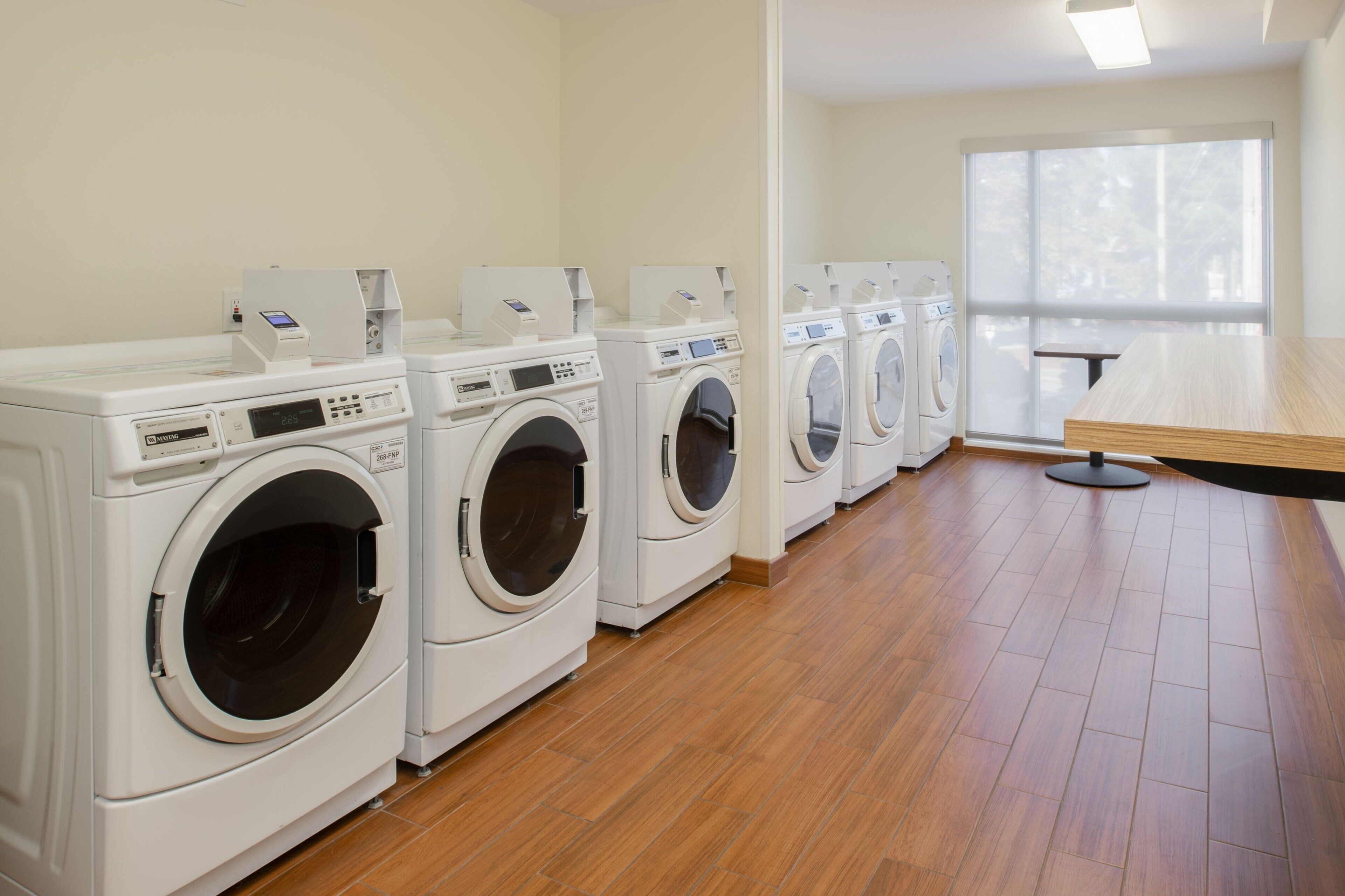 A laundry room with multiple washing machines and dryers, wooden flooring, and a large window providing natural light.