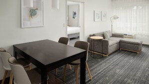 A modern hotel room with a dining area, a gray sofa, a round side table, and a large window with sheer curtains.