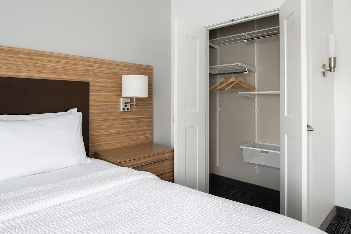 Siisti hotellihuone, jossa on huolitellusti pedattu sänky, puinen pääty, yöpöytälamppu, vaatehuone hyllyineen ja henkareineen sekä ovessa oleva kahva.