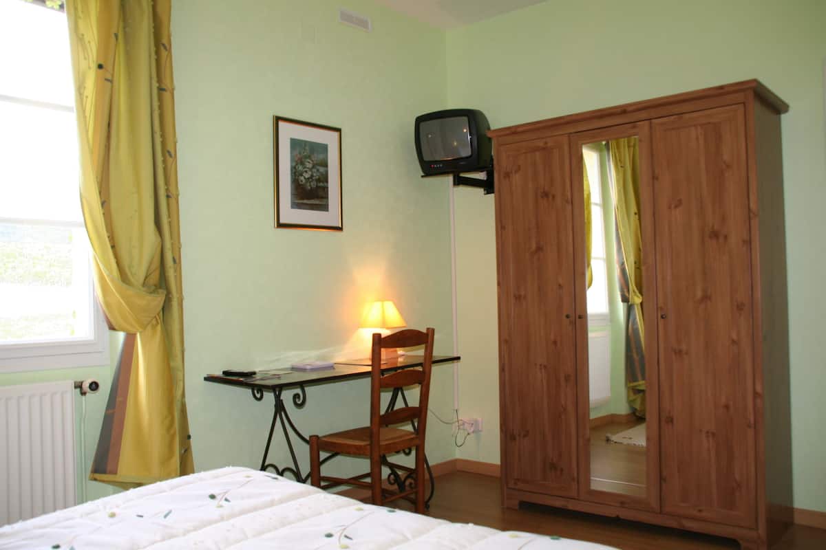 A bedroom with a bed, a desk with a lamp, a chair, a wardrobe, and a television on the wall.