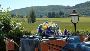Un área de comedor al aire libre con sillas de mimbre y una mesa para dos, con vistas a un paisaje pintoresco con colinas y un edificio distante.