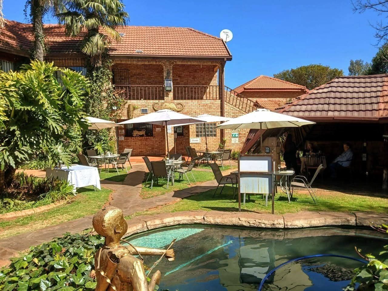 A pool with a small waterfall feature, surrounded by outdoor seating and umbrellas, with a brick building in the background.