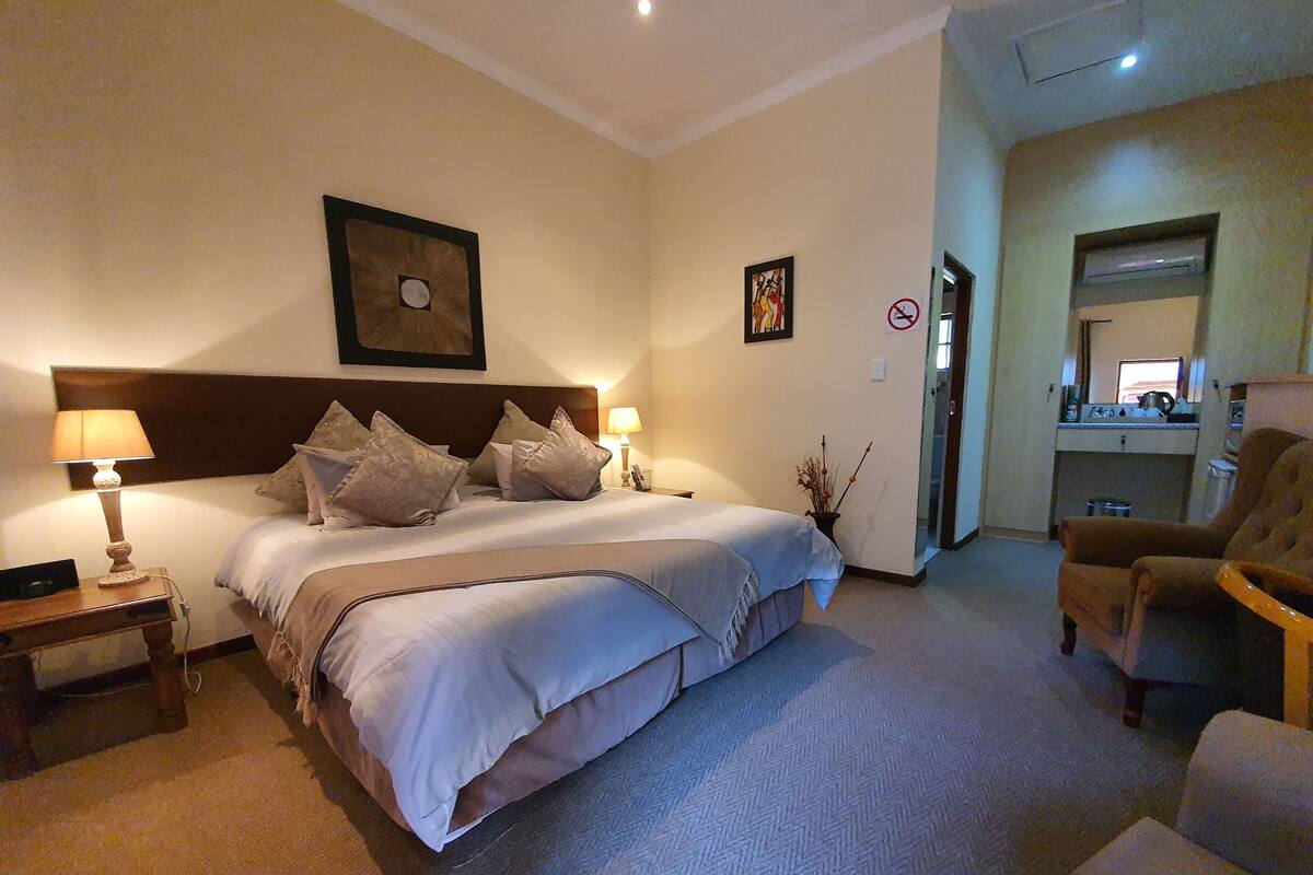 Ein Hotelzimmer mit einem großen Bett, Nachttischlampen, einem kleinen Tisch, einem Sessel und einer Küchenzeile im Hintergrund.
