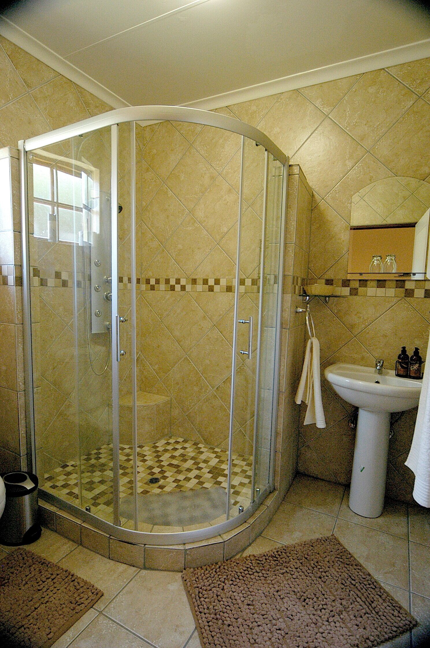 A corner shower with a glass door, beige tiled walls, a sink, and a towel rack.