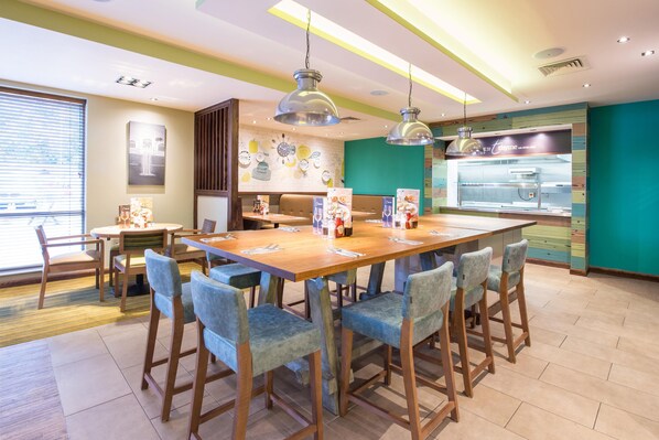 Un restaurant avec des tables et des chaises en bois, un espace cuisine et un mur orné d’horloges et d’ustensiles de cuisine.