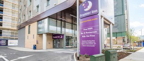 De ingang van een Premier Inn hotel met een bord, een parkeerplaats en een modern gebouw op de achtergrond.