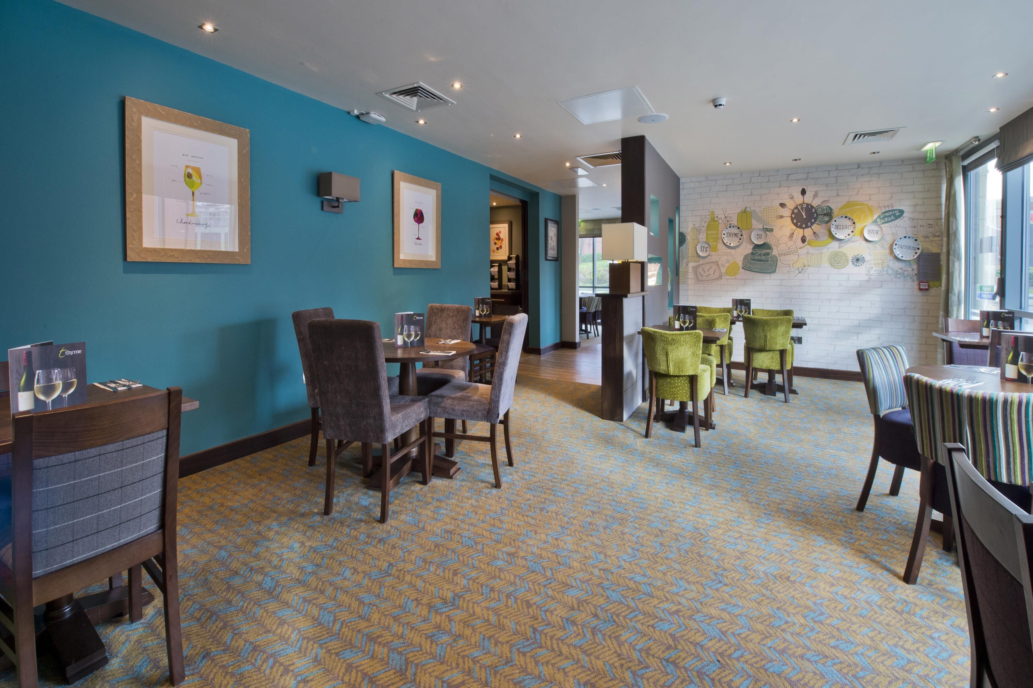 Ein Hotel-Speisesaal mit mehreren Tischen und Stühlen, gerahmten Kunstwerken an den Wänden und einem gemusterten Teppich.