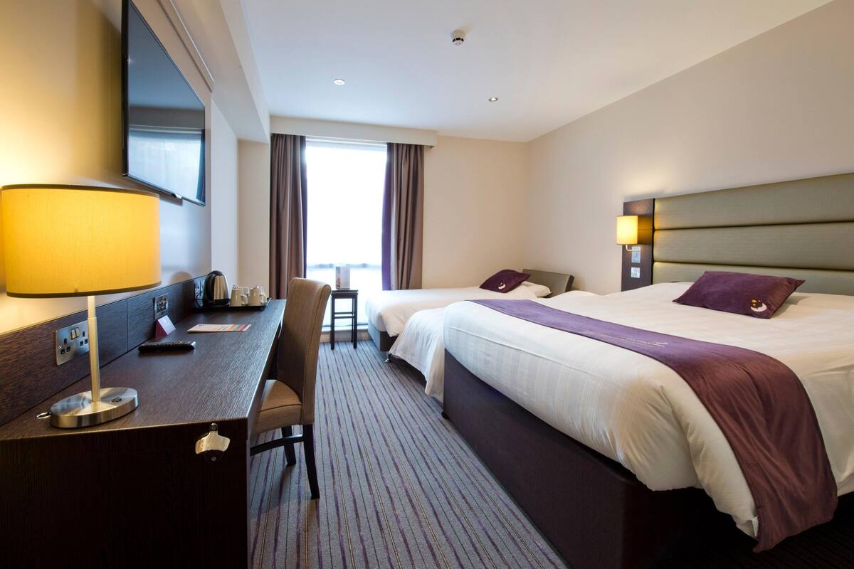 A hotel room with a desk, two beds, a chair, a TV, and a window with curtains.