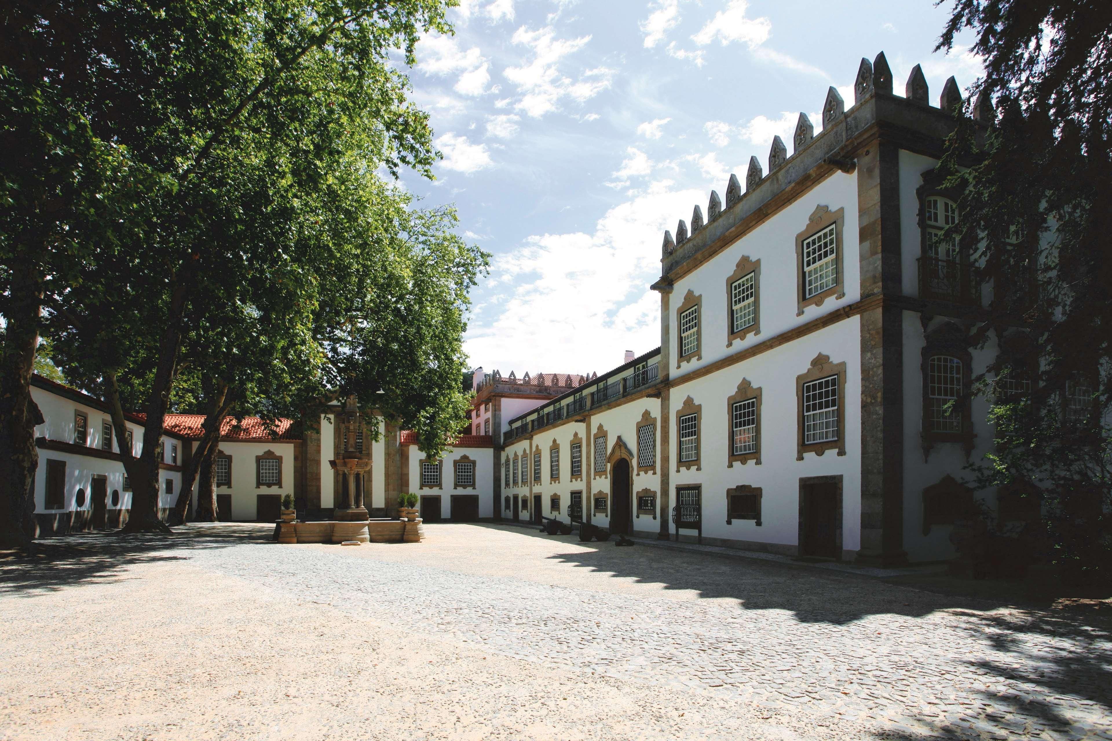 A courtyard with a cobblestone ground, white buildings with arched windows, and a large tree providing shade.