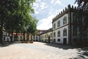 Um pátio com chão de pedra, edifícios brancos com janelas em arco e uma grande árvore oferecendo sombra.