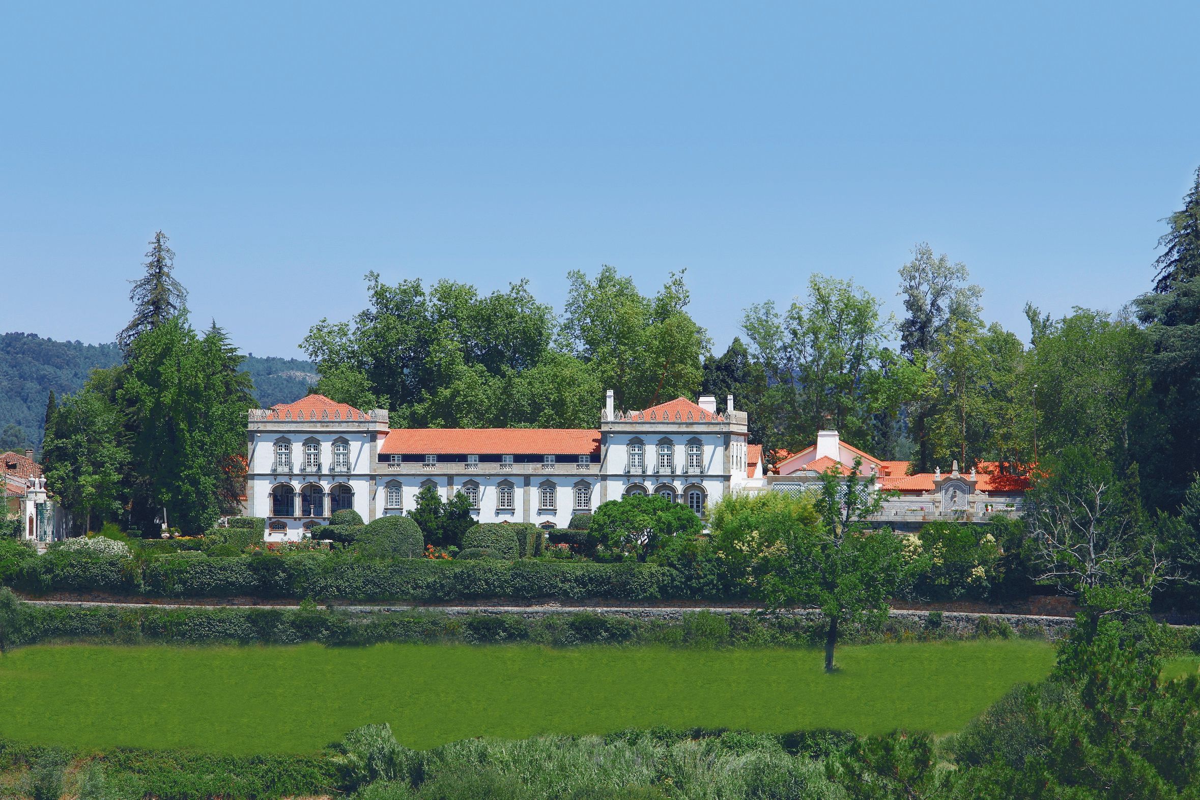 A large, historic building with a red-tiled roof, surrounded by lush greenery and a well-maintained lawn.