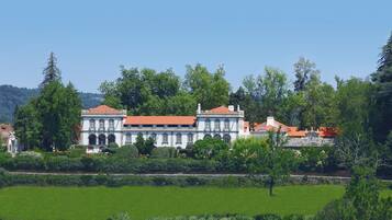 A large, historic building with a red-tiled roof, surrounded by lush greenery and a well-maintained lawn.