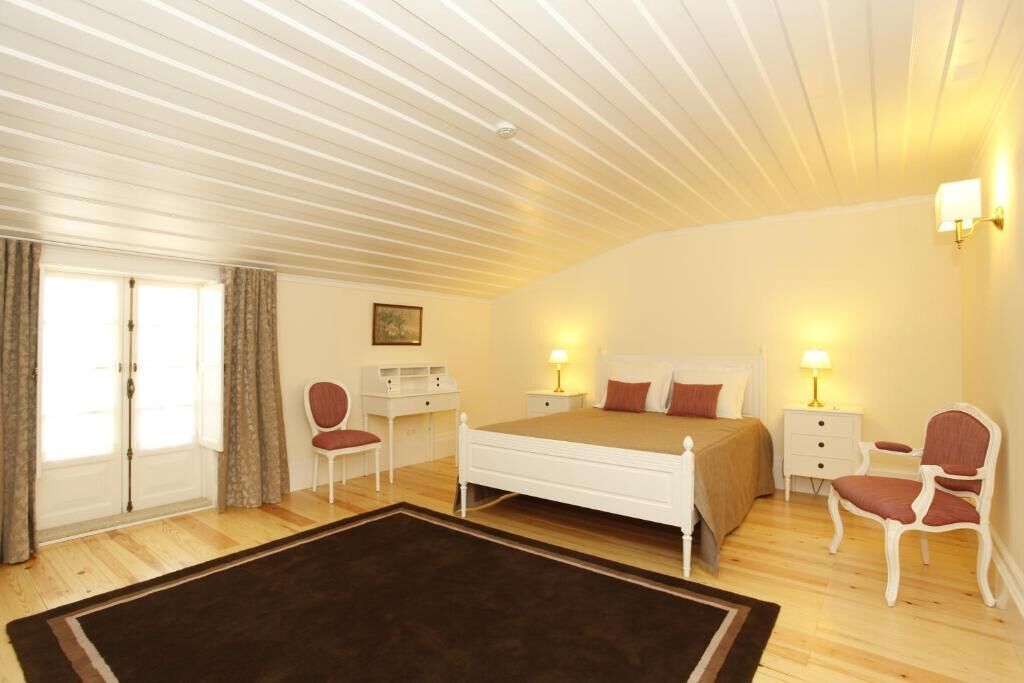 Um quarto com cama, mesas de cabeceira, uma cadeira, escrivaninha e janela com cortinas.