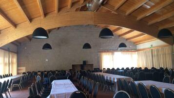 Un gran salón con techos altos de madera, varias mesas con sillas y un escenario al frente.