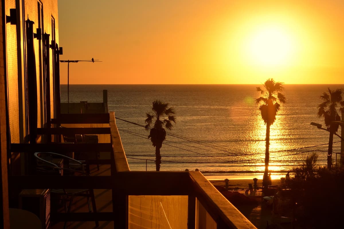 A balcony with a view of the sunset over the ocean, featuring palm trees and a wooden railing.