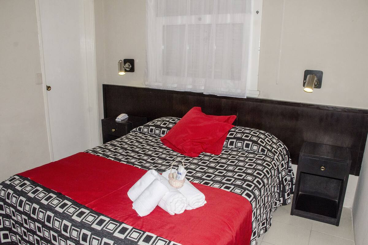 A hotel room with a bed featuring a geometric patterned bedspread, a red throw pillow, and neatly folded white towels. There is a bedside table with a phone and a small decorative item.