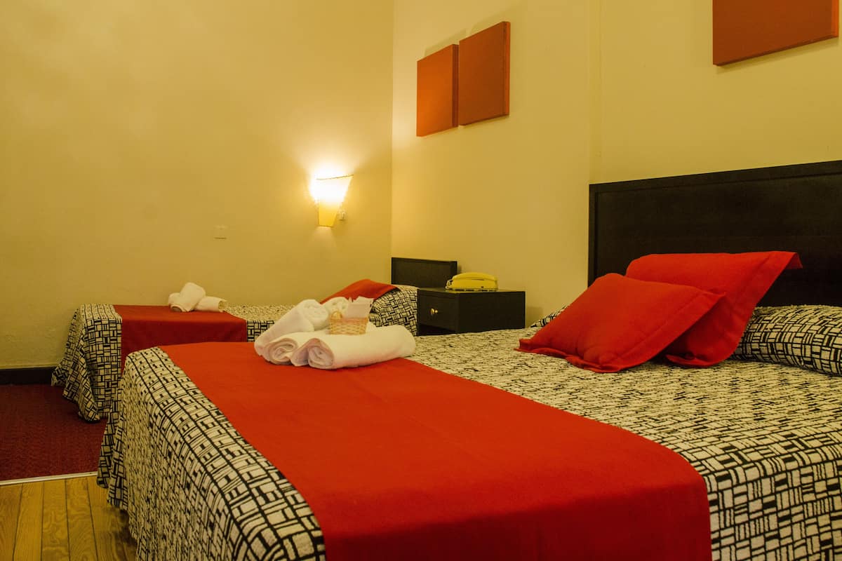 A hotel room with two beds, red and black patterned bedding, a nightstand with a telephone, and a wall-mounted light.