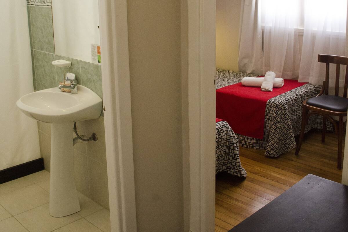 A bathroom with a sink and a shower. There is a bed with a red blanket and a chair.
