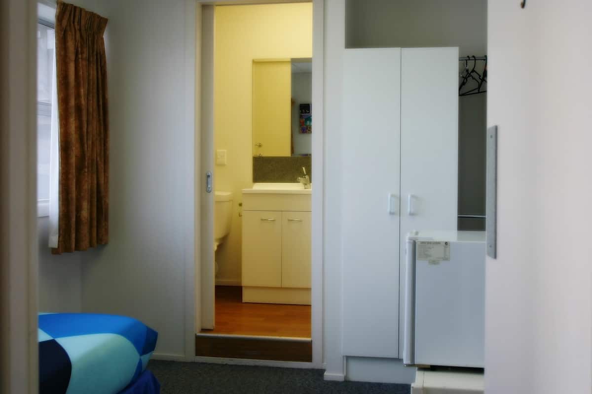 Quarto compacto com uma cama, um guarda-roupa e uma porta que dá para uma cozinha.